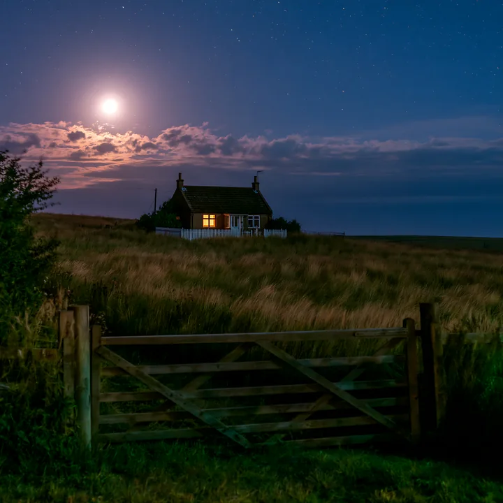 A house sits alone in a field, illuminated by the light of a full moon and stars.