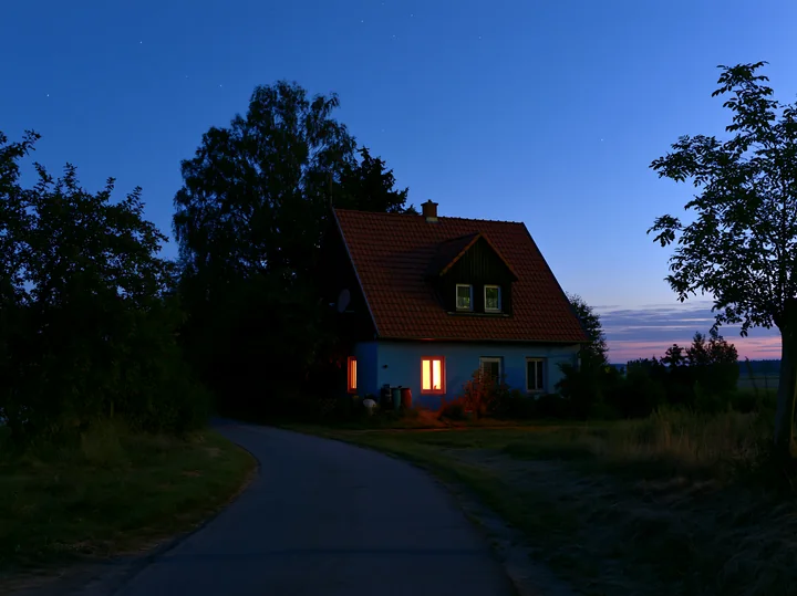 A house with warm light glowing from its windows stands beside a winding road, surrounded by trees and grass, as stars twinkle in the blue hour sky.
