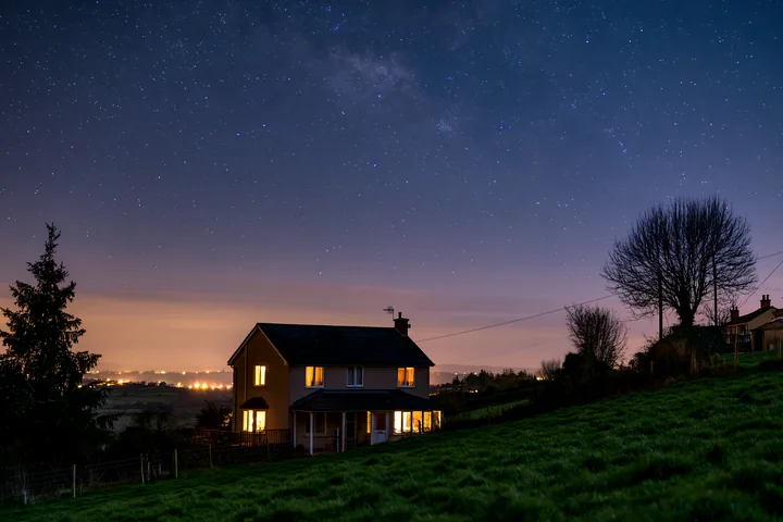 A house with warm lights shining from the windows stands in the countryside under a starry sky.
