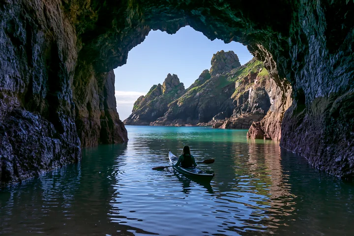 A kayaker paddles through a sea cave, with a clear view of a rocky coastline outside.