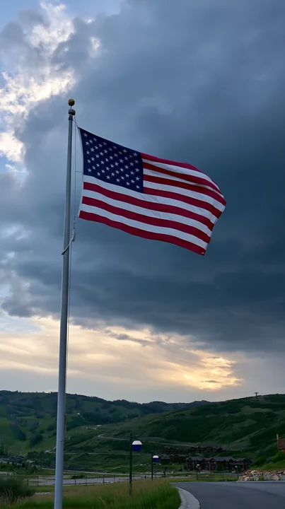 A large American flag is flying in strong wind on a pole, set against a backdrop of dramatic clouds.