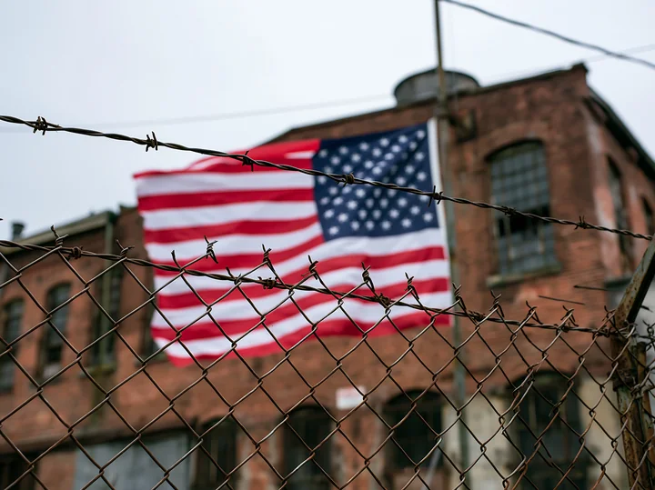 A large American flag is visible behind a barbed wire fence in an industrial area.