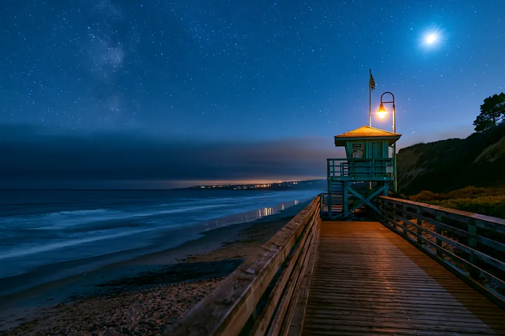 A lifeguard tower stands at the end of a coastal boardwalk under a starry night sky.