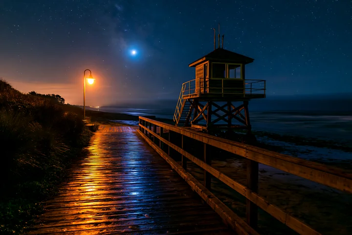A lifeguard tower stands beside a wooden boardwalk under a starry night sky.