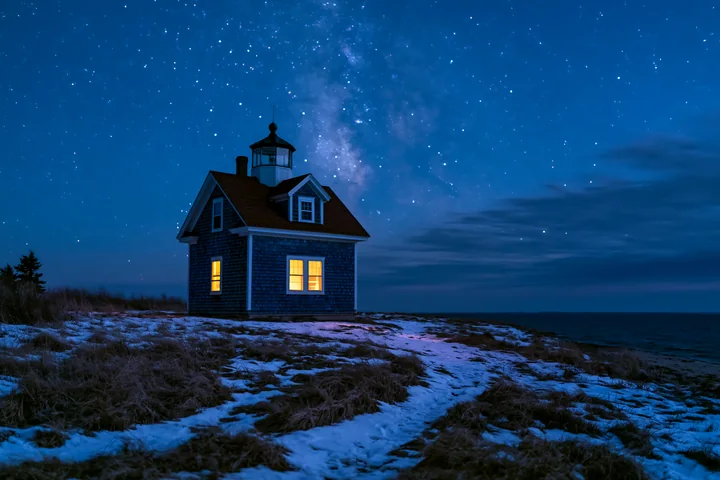 A lighthouse house stands on a grassy area near the shore under a starry sky.