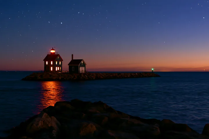 A lighthouse house under stars stands on a rocky jetty at dusk, with a calm sea reflecting the lights.