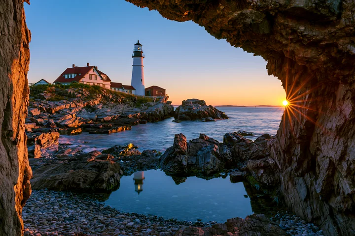 A lighthouse near a rocky tide pool stands tall as the sun sets on the horizon.