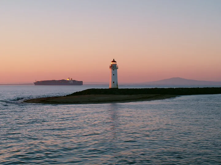 A lighthouse stands on a rocky point as a cargo ship travels in the water at sunset.