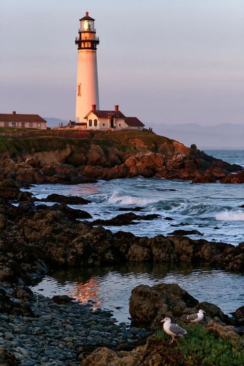 A lighthouse stands tall near a rocky tide pool as waves crash against the shore.