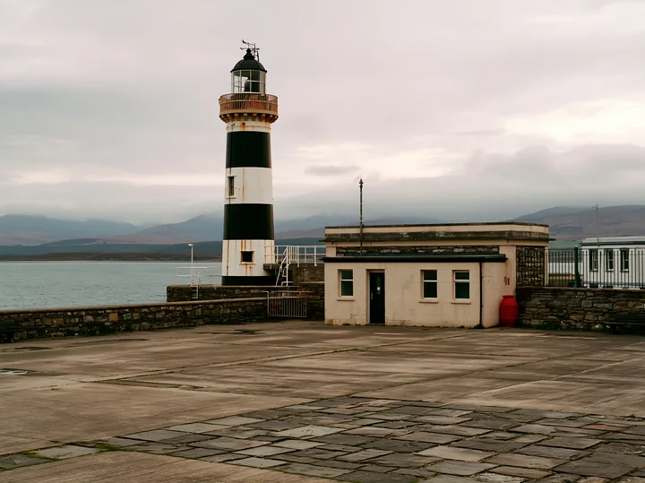 A lighthouse with black and white stripes stands near a simple coastal building, surrounded by calm waters and cloudy skies.