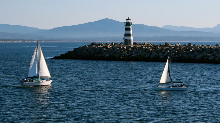 A lighthouse with black and white stripes stands on a rocky jetty as sailboats glide through the water, with mountains visible in the background during clear we