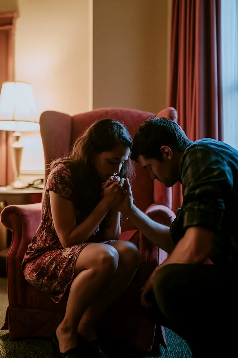 A man gently kisses a woman's hand while they sit together in a warmly lit living room.