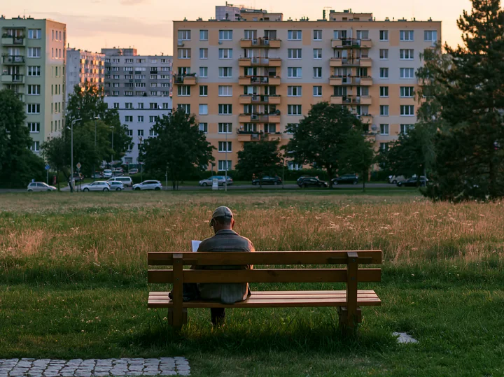 A man sits on a wooden bench in an urban meadow among buildings, reading as the sun sets behind tall residential structures.
