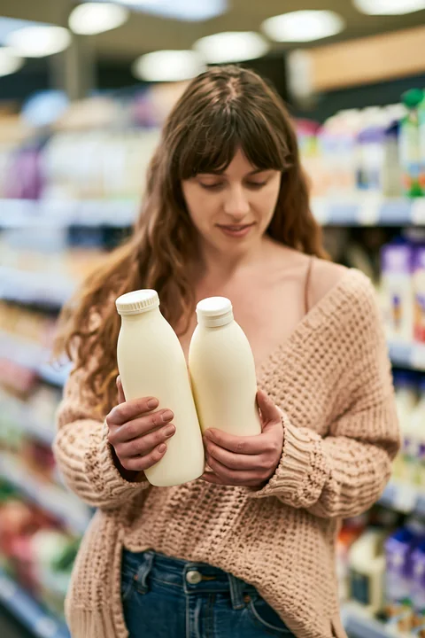 A market customer comparing different local milk bottles holds two bottles while standing in a grocery aisle.