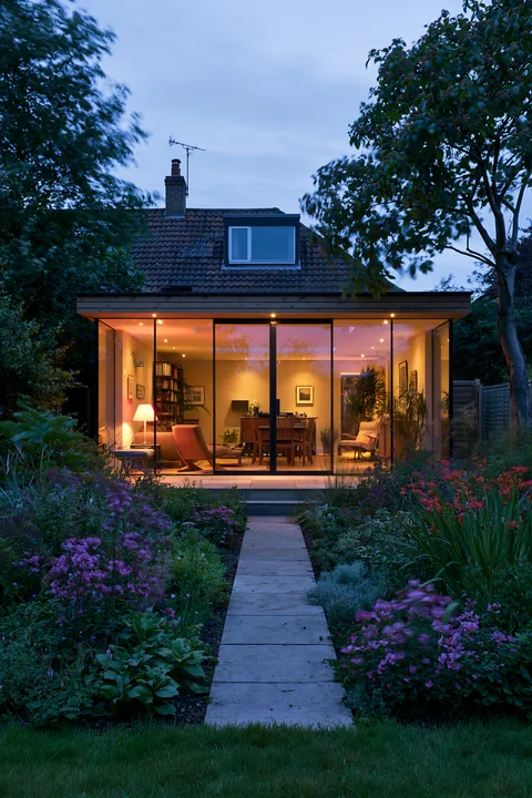 A modern empty garden house with glowing glass walls showcases a well-lit interior filled with furniture.