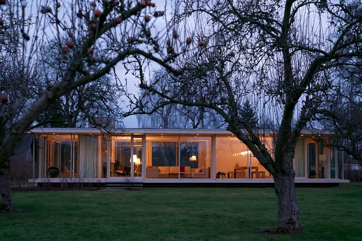 A modern empty garden house with glowing glass walls surrounded by trees as twilight approaches.