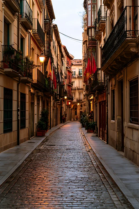A narrow cobblestone street is lined with buildings featuring balconies and decorative flags.