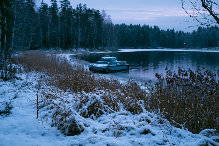 A panzer tank in winter snow moves along the shoreline of a frozen lake, with tall grasses and snow-covered ground in the background.
