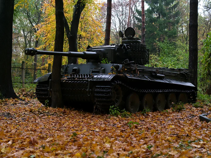 A panzer tank is parked among trees in an autumn forest, surrounded by orange and yellow leaves on the ground.