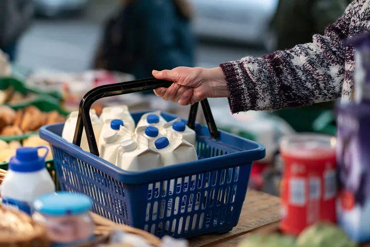 A person holds a blue shopping basket that contains milk cartons while standing at a market.