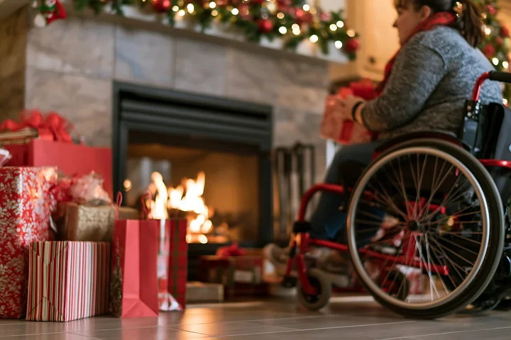 A person in a wheelchair sits beside a warm fireplace surrounded by colorful holiday shopping bags and presents.
