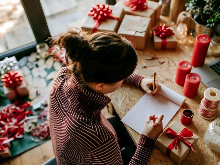 A person is focused on writing a gift list at a desk surrounded by holiday decorations and wrapped gifts.