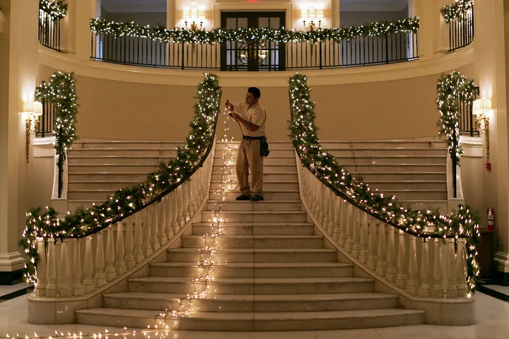 A person is hanging string lights along a grand staircase decorated with greenery.