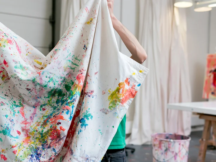 A person lifts a piece of cotton fabric covered with colorful paint splatter in an art studio.