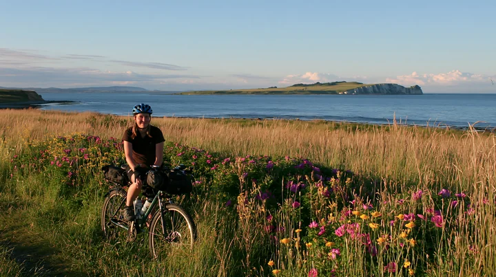 A person rides a bicycle along a path lined with flowers and tall grass, overlooking the ocean and white cliffs on an island in the distance during daylight.