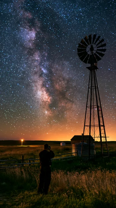 A person using camera under Milky Way stands near a windmill in an open field.