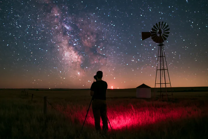 A person using camera under Milky Way stands on a grassy field beside a windmill.
