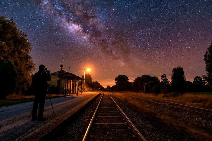 A person using camera under Milky Way stands on a quiet train station platform.