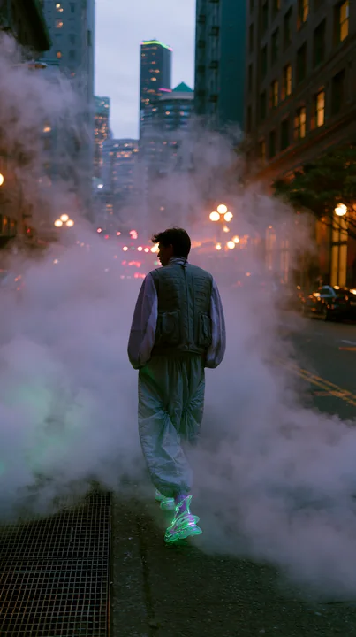 A person walks down a city street enveloped in fog as lights illuminate the buildings.