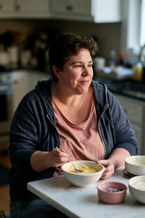 A person with limited hand mobility using utensils sits at a kitchen table, enjoying a bowl of soup.