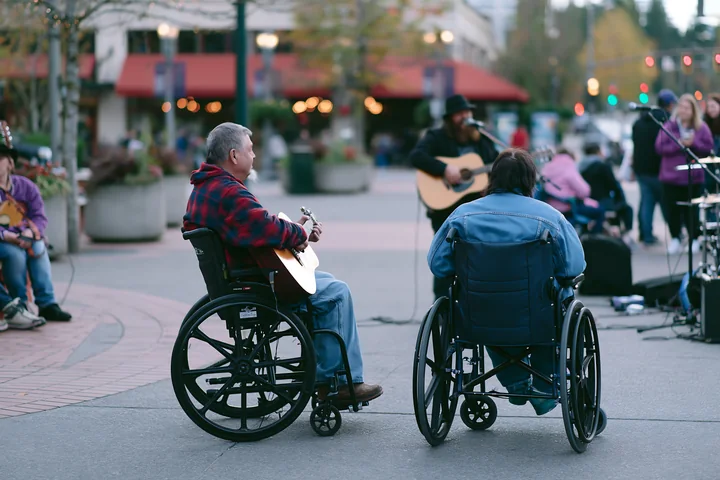 A person with muscular dystrophy using a wheelchair enjoys a live music performance in a busy outdoor setting.