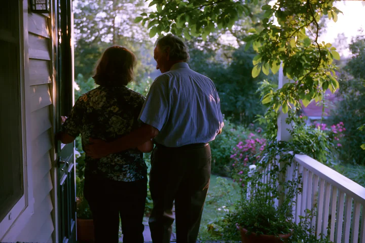 A person with Parkinson's disease receiving support stands beside another individual on a porch in a garden.
