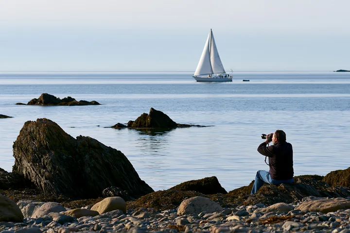 A photographer photographing a sailboat at sea from a rocky shore.