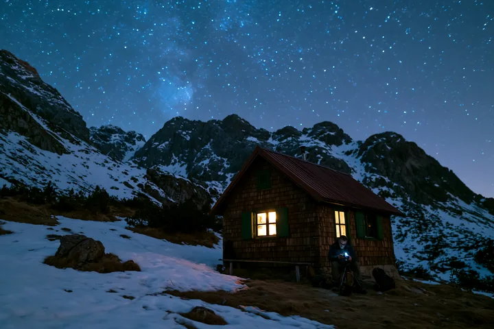 A photographer under stars in the mountains sets up a camera outside a rustic cabin.