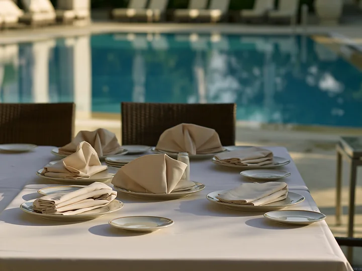 A poolside buffet table is arranged with plates and folded napkins beside a clear blue swimming pool.