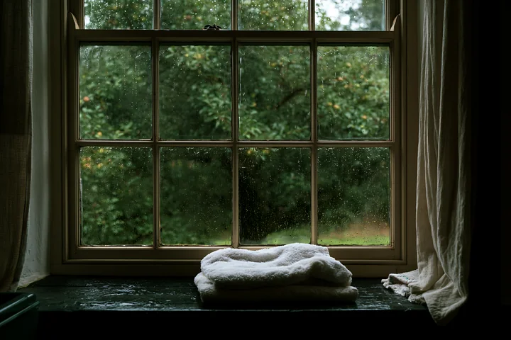 A rain-soaked wooden window in a rural home shows blurred greenery outside.