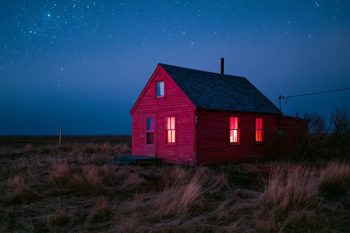 A red cabin stands alone in a grassy field as stars fill the night sky.
