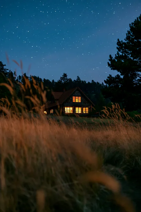 A remote mountain home under stars stands out in the night, with warm light glowing from its windows.
