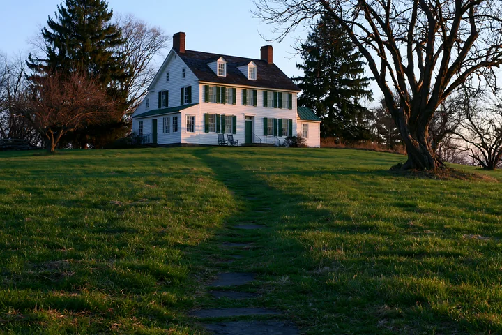 A restored farmhouse exterior stands on a hill, surrounded by green grass and large trees.
