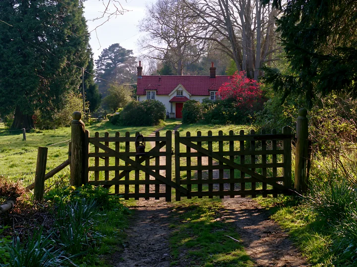 A rural cottage is seen through a wooden gate along a dirt path.