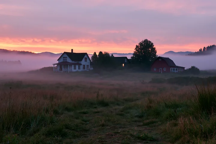 A rural farmhouse in morning fog is seen against a colorful sky.