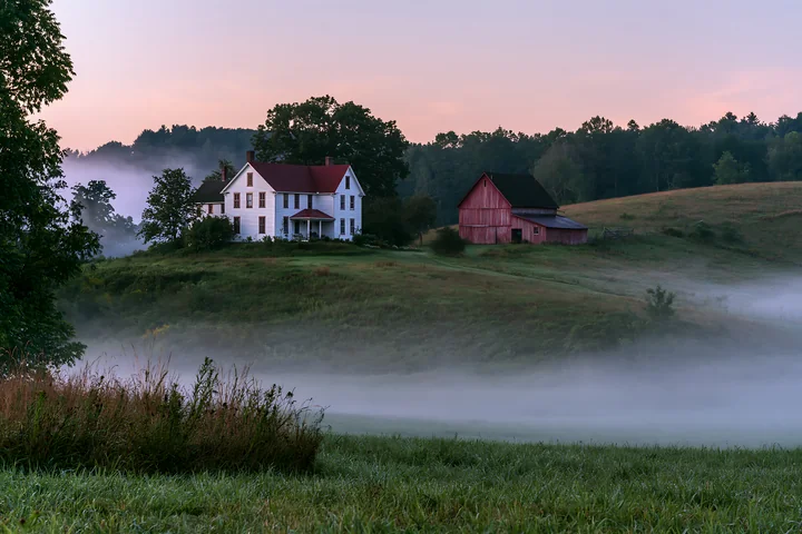 A rural farmhouse in morning fog sits on a hill, surrounded by fields and trees.