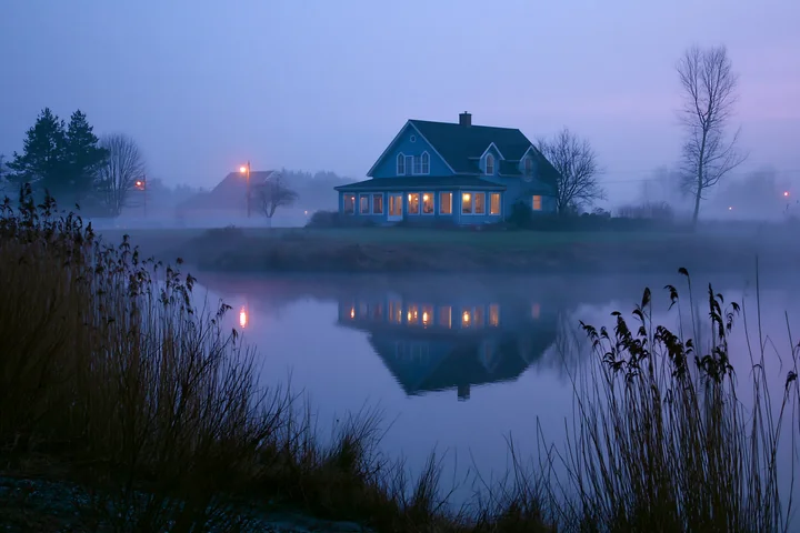 A rural farmhouse in morning fog stands by a quiet lake.