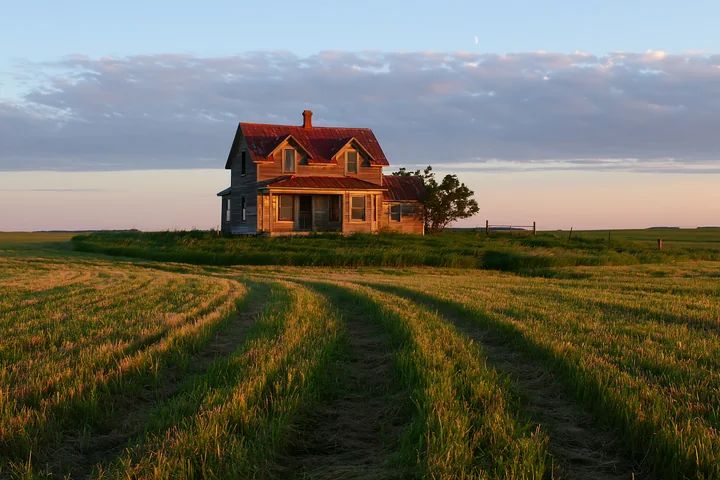 A rustic farmhouse stands in golden light, surrounded by open fields and gentle hills.