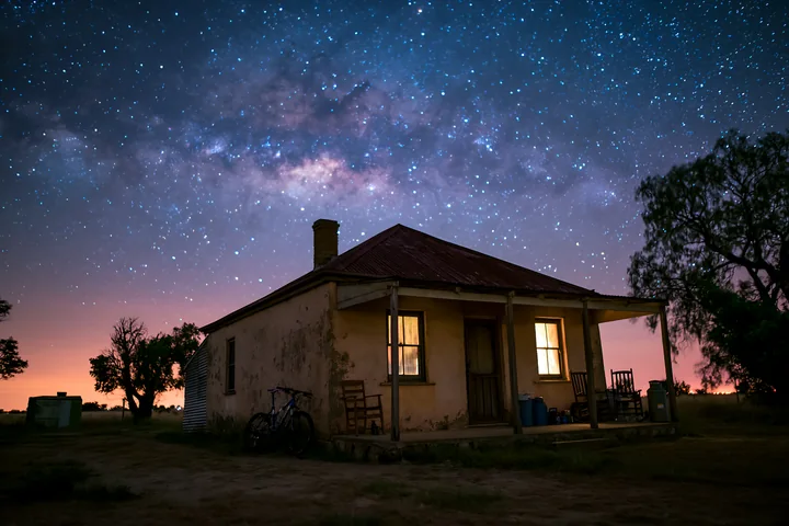 A rustic farmhouse stands under a bright night sky filled with stars and the Milky Way.