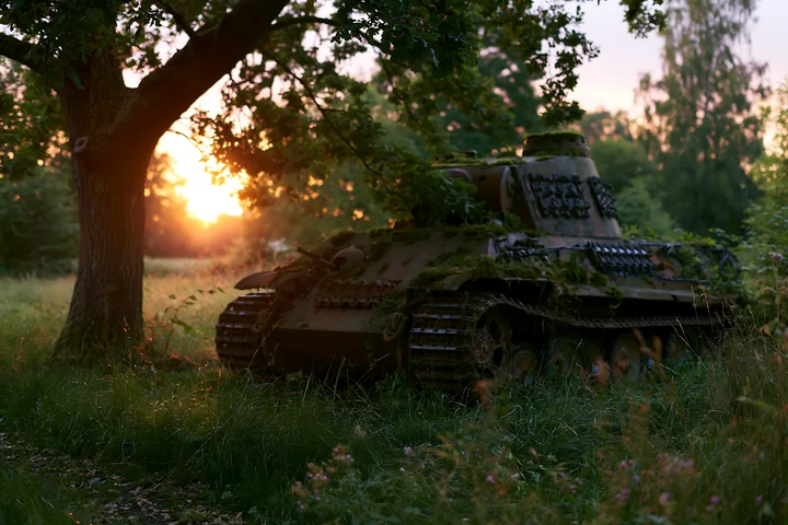 A rusty tank sits in a grassy field as the sun sets behind it, creating a warm glow.